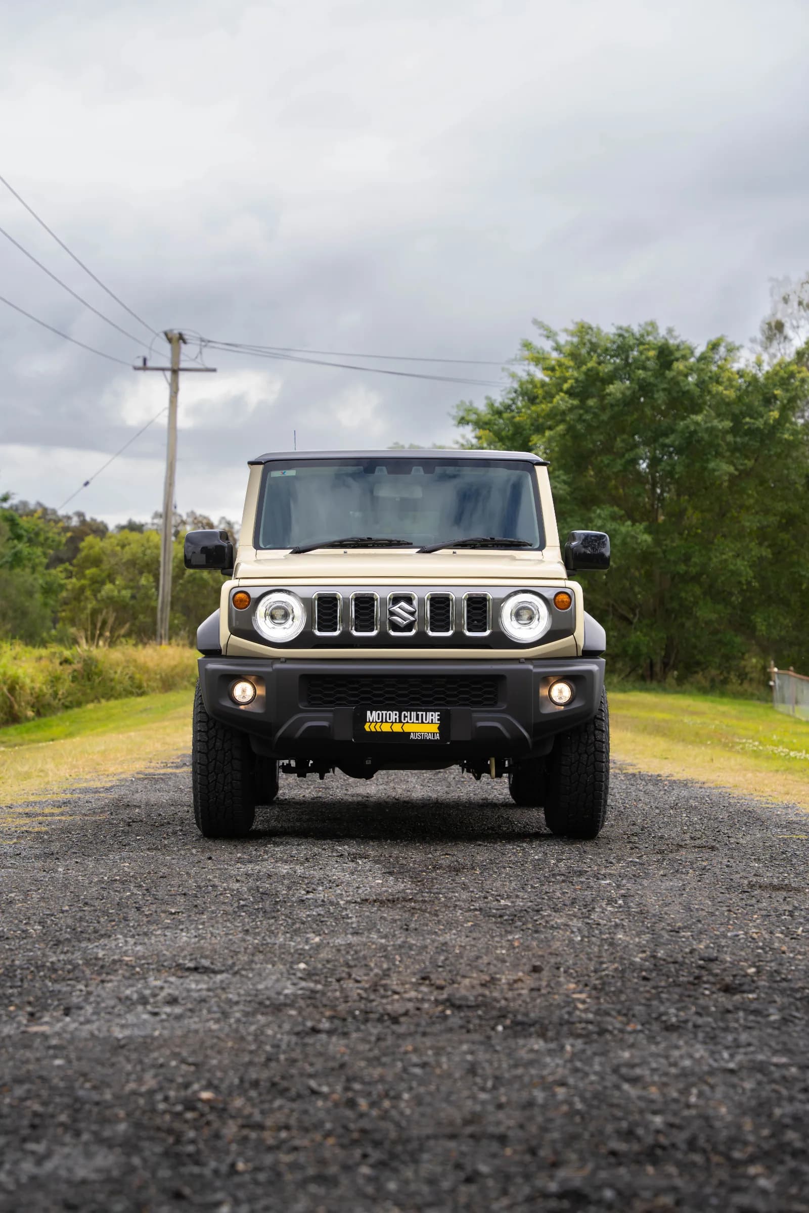 Suzuki Jimny XL in sandy beige faces forward on a gravel road with overcast sky.