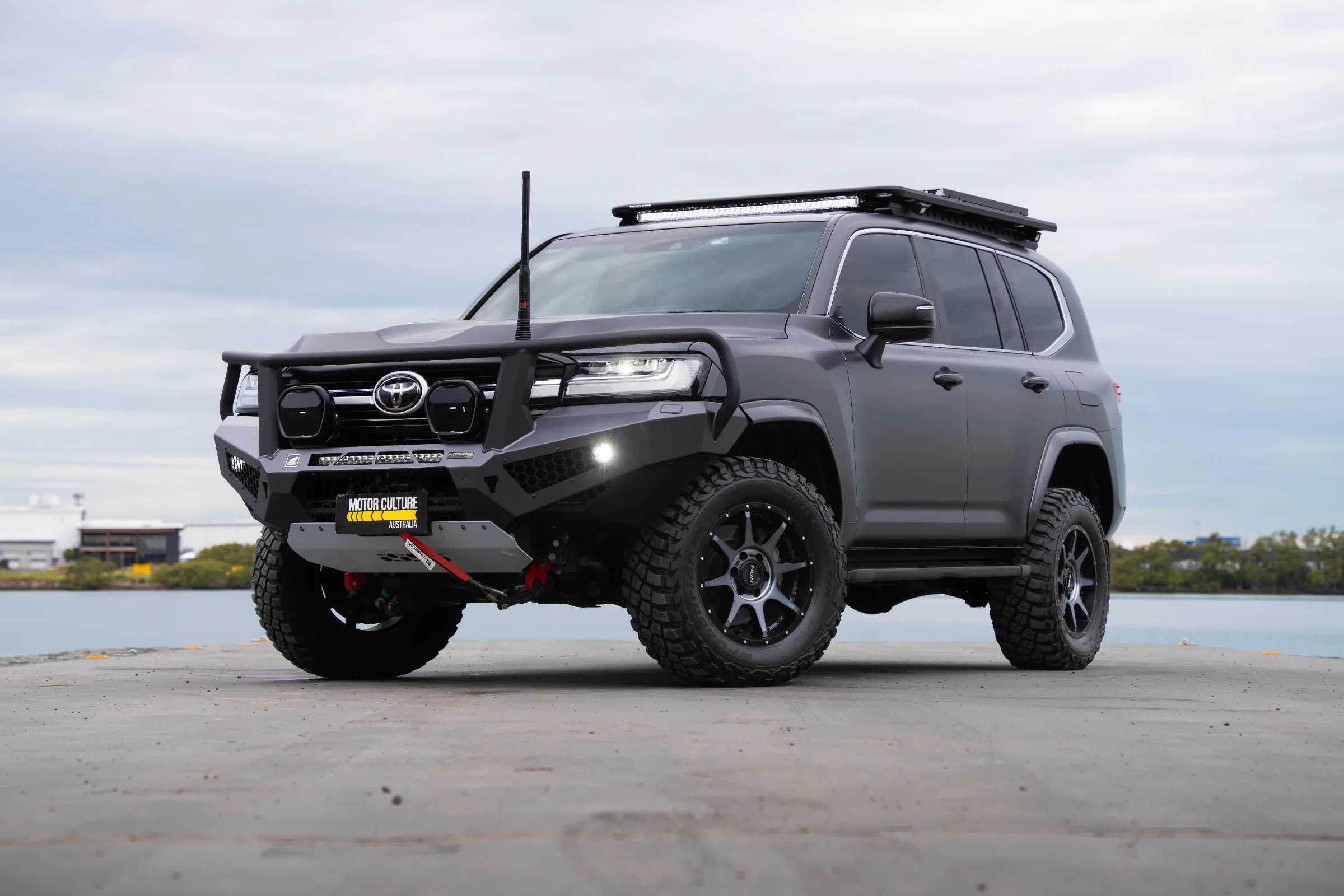 Matte grey Toyota LandCruiser 300 Series Sahara with bull bar, roof rack and light bar parked on a concrete boat ramp